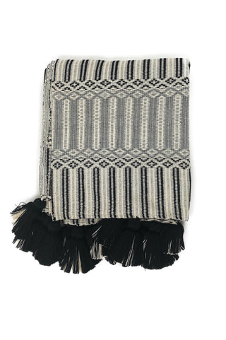 Volcanic Sands Throw by Del Palomar. Handmade in Guatemala. Handwoven authentic Guatemalan fabric throw, featuring an intricately woven textured pattern and adorned with black and white tassels. Handwoven on a traditional footloom by local artisans in Chimaltenango, Guatemala. Color black and white. 100% cotton.